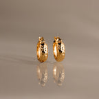Bold Hammered Huggies in 18K Gold with a textured finish and abstract black designs are displayed on a reflective, neutral-colored surface.