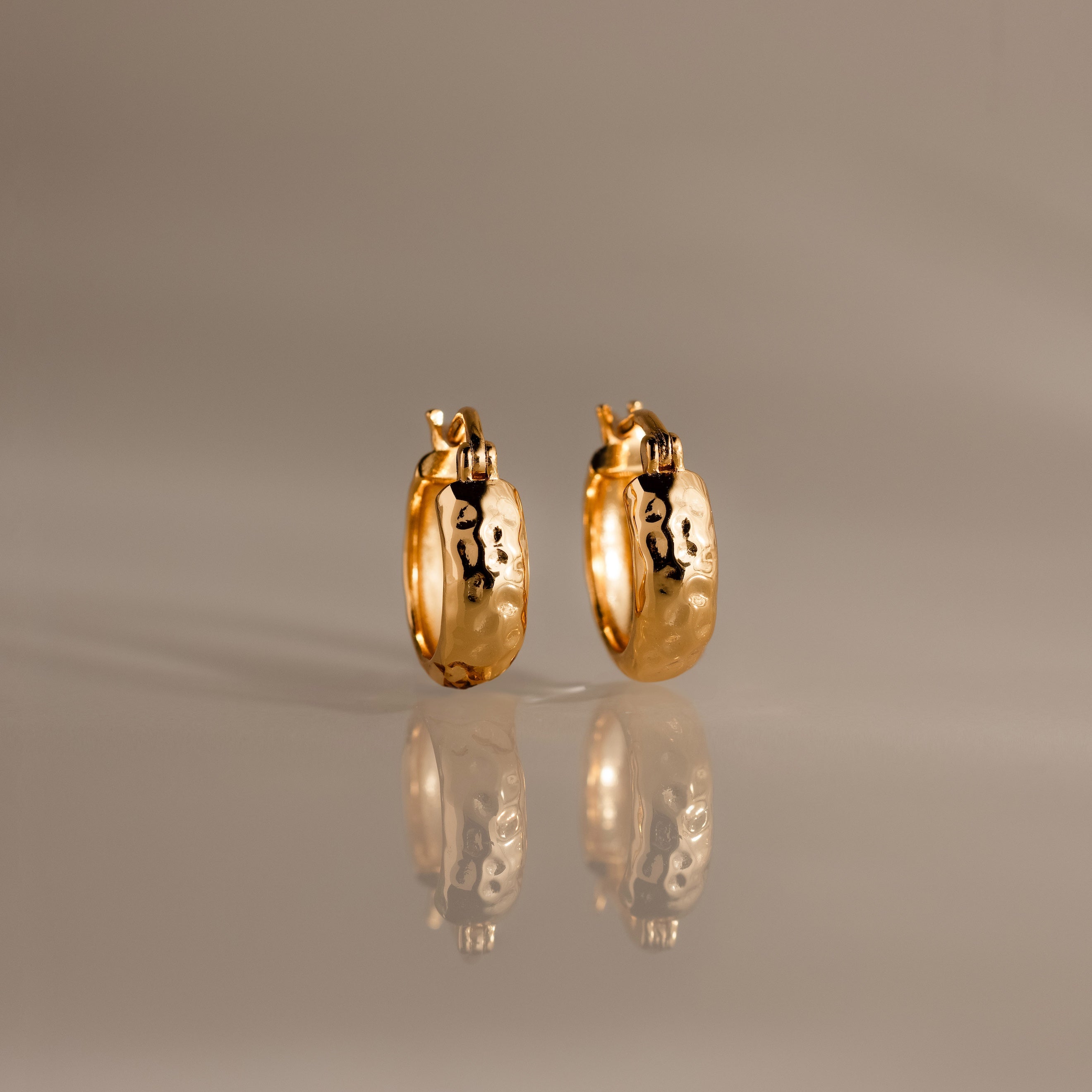 Bold Hammered Huggies in 18K Gold with a textured finish and abstract black designs are displayed on a reflective, neutral-colored surface.