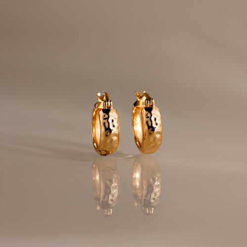 Bold Hammered Huggies in 18K Gold with a textured finish and abstract black designs are displayed on a reflective, neutral-colored surface.