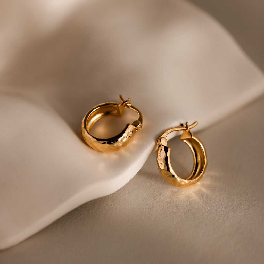 The Bold Hammered Huggies in 18K Gold, featuring a textured finish, rest on soft cream fabric, exemplifying the elegance of minimal design jewelry.