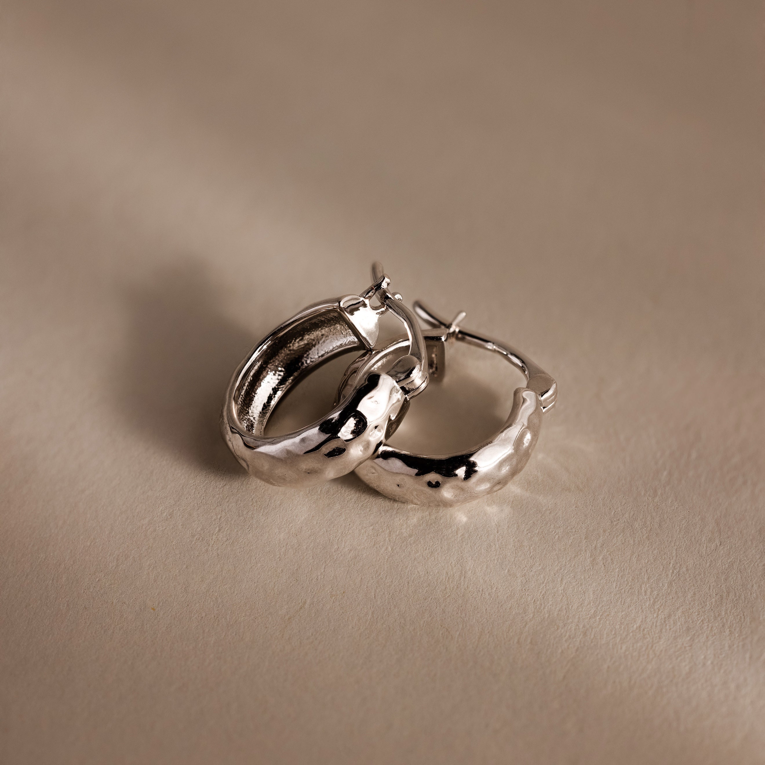 The Bold Hammered Huggies in Silver, featuring a textured finish, rest on a soft neutral surface—an ideal choice for fans of minimal design jewelry.