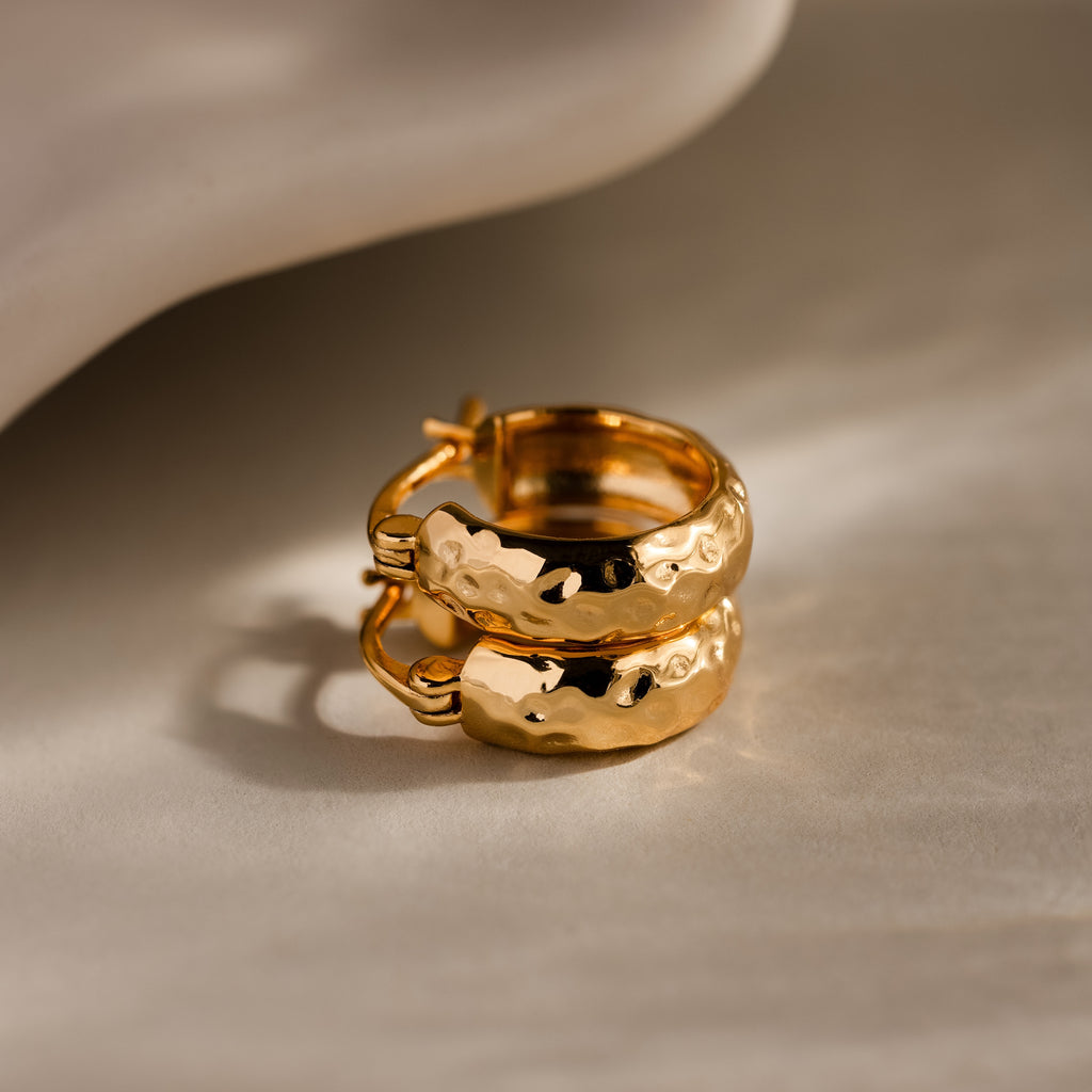 Two Bold Hammered Huggies in 18K Gold with a textured finish are displayed on a light, textured surface with soft shadows, highlighting their minimal design.