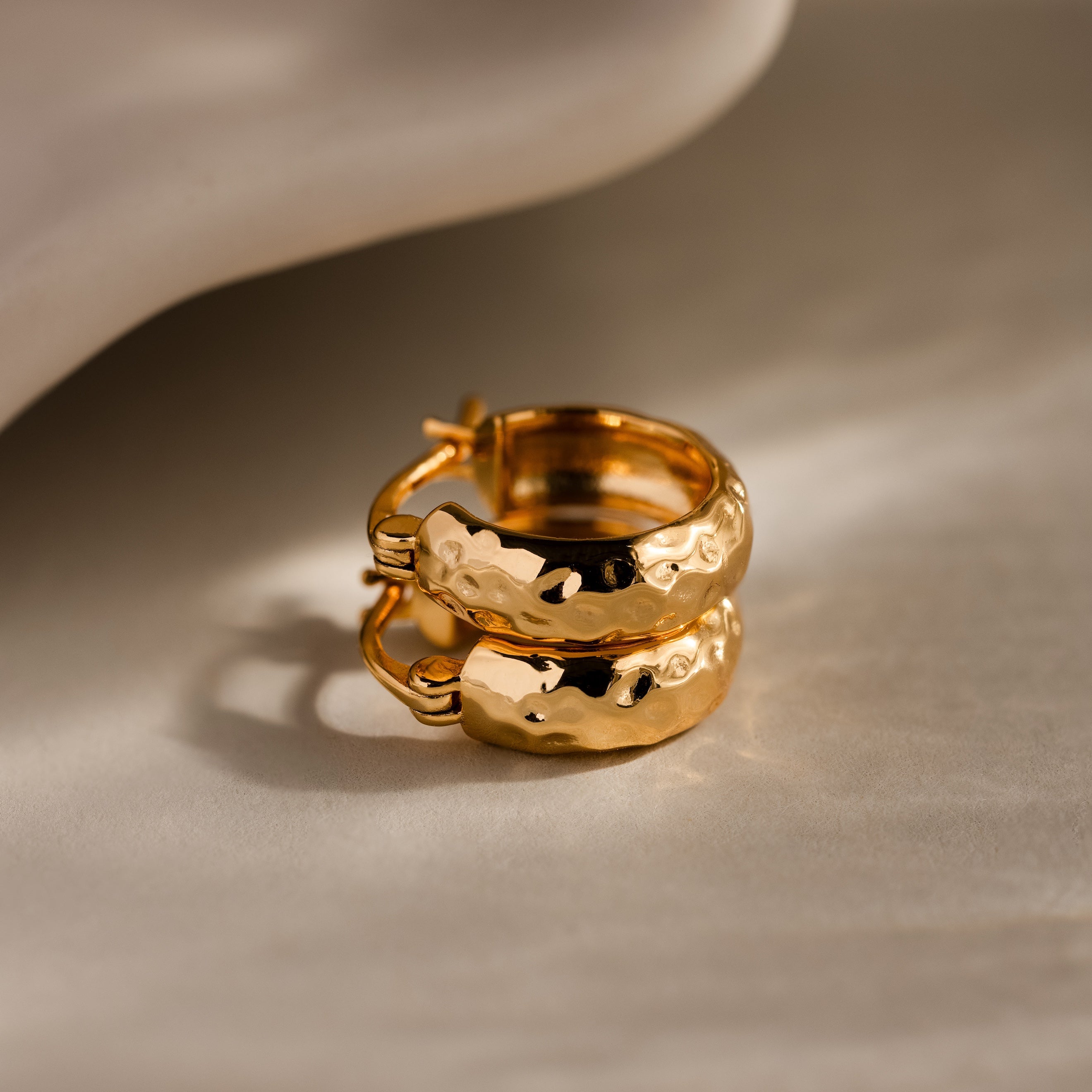 Two Bold Hammered Huggies in 18K Gold with a textured finish are displayed on a light, textured surface with soft shadows, highlighting their minimal design.