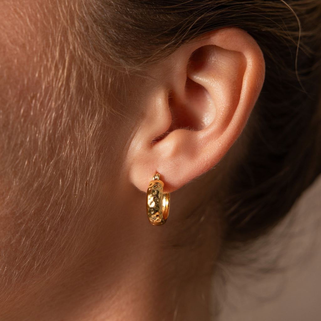 Close-up of a person’s ear wearing Bold Hammered Huggies in 18K Gold, highlighting the beauty of minimal design jewelry with a textured gold finish.