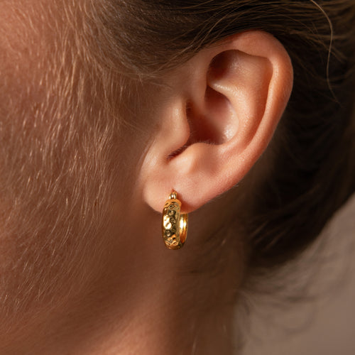 Close-up of a person’s ear wearing Bold Hammered Huggies in 18K Gold, highlighting the beauty of minimal design jewelry with a textured gold finish.