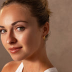 Woman with blonde hair in a bun, wearing Bold Hammered Huggies in 18K Gold and a white top, smiling softly at the camera.