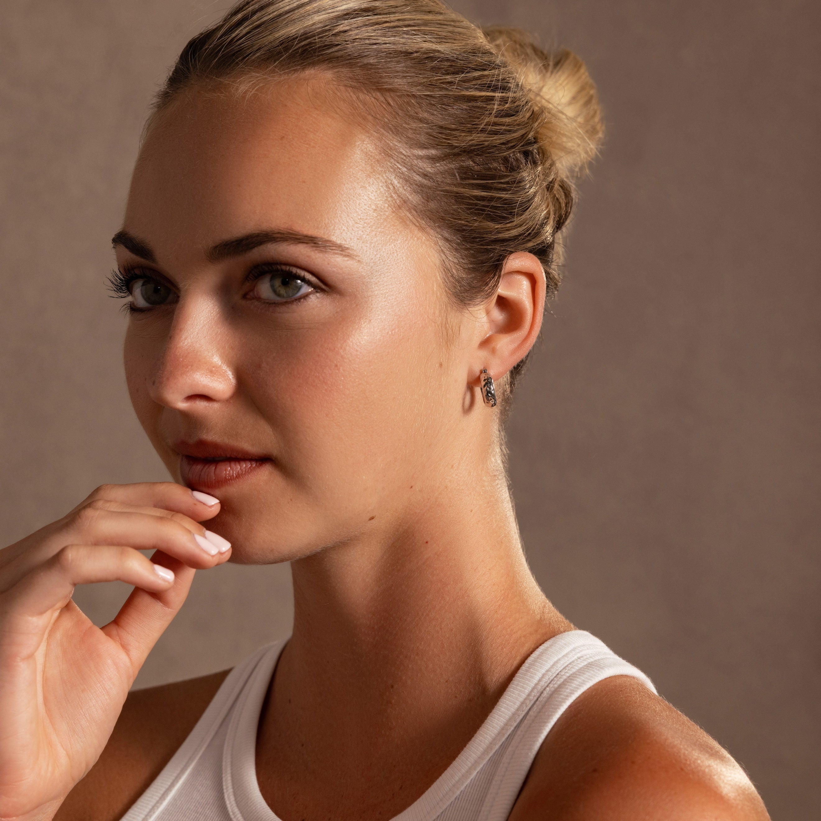 A woman with blonde hair in a bun, wearing a white tank top and the Bold Hammered Huggies, gazes thoughtfully to the side.