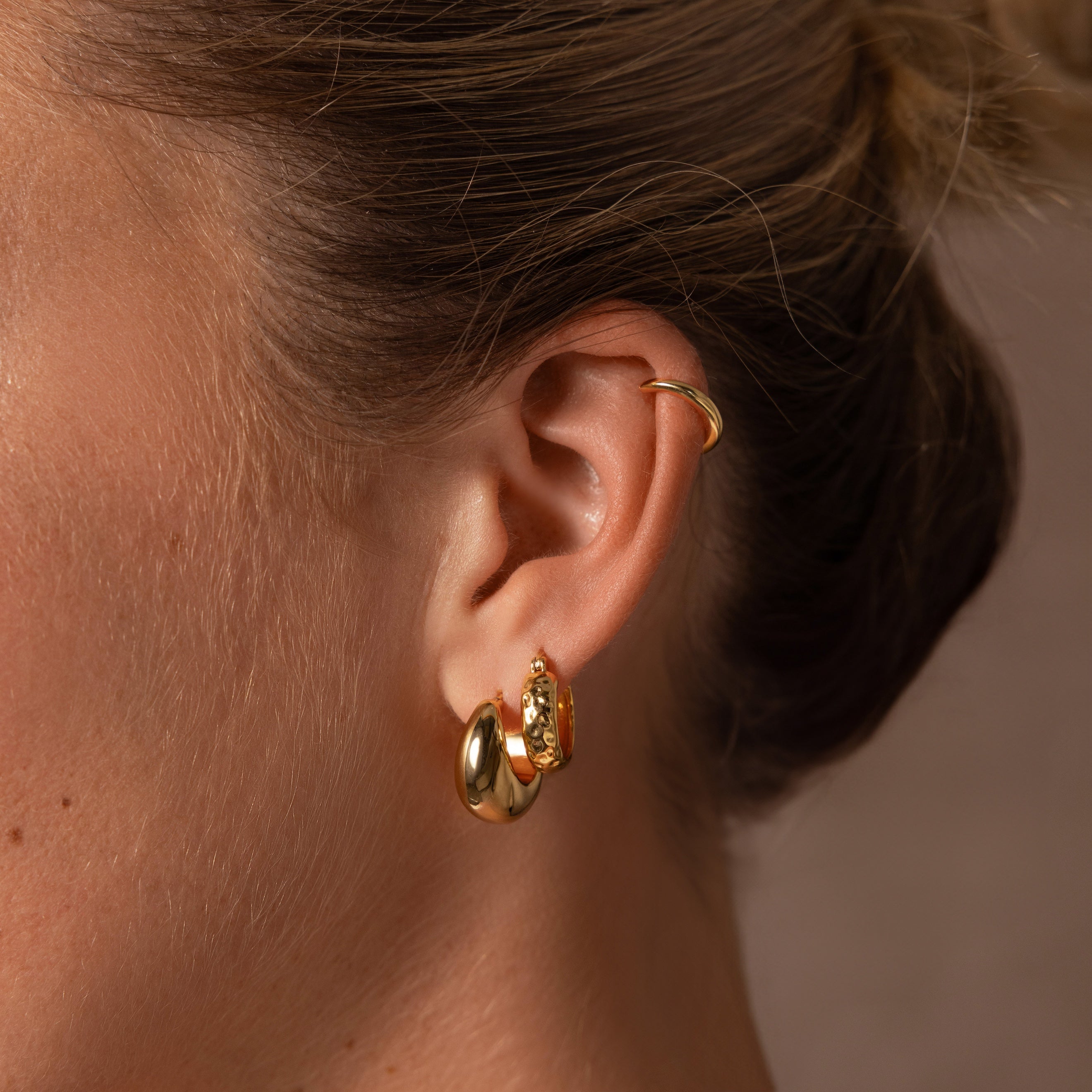 A close-up of a womans ear adorned with three gold earrings, including a hoop and the Bold Hammered Huggies in 18K Gold, highlighting minimal design with a stylish, textured finish.