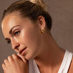 A woman with blonde hair in a bun, wearing Bold Hammered Huggies in 18K Gold, rests her chin on her hand and wears a white top.