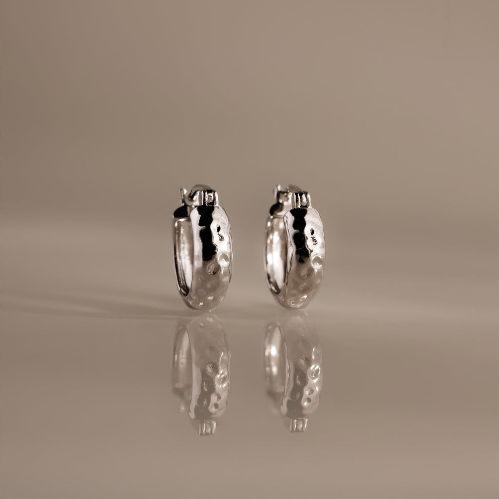 Bold Hammered Huggies in Silver with a textured finish stand upright on a reflective surface, highlighting their minimal design against a neutral background.