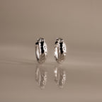 Bold Hammered Huggies in Silver with a textured finish stand upright on a reflective surface, highlighting their minimal design against a neutral background.