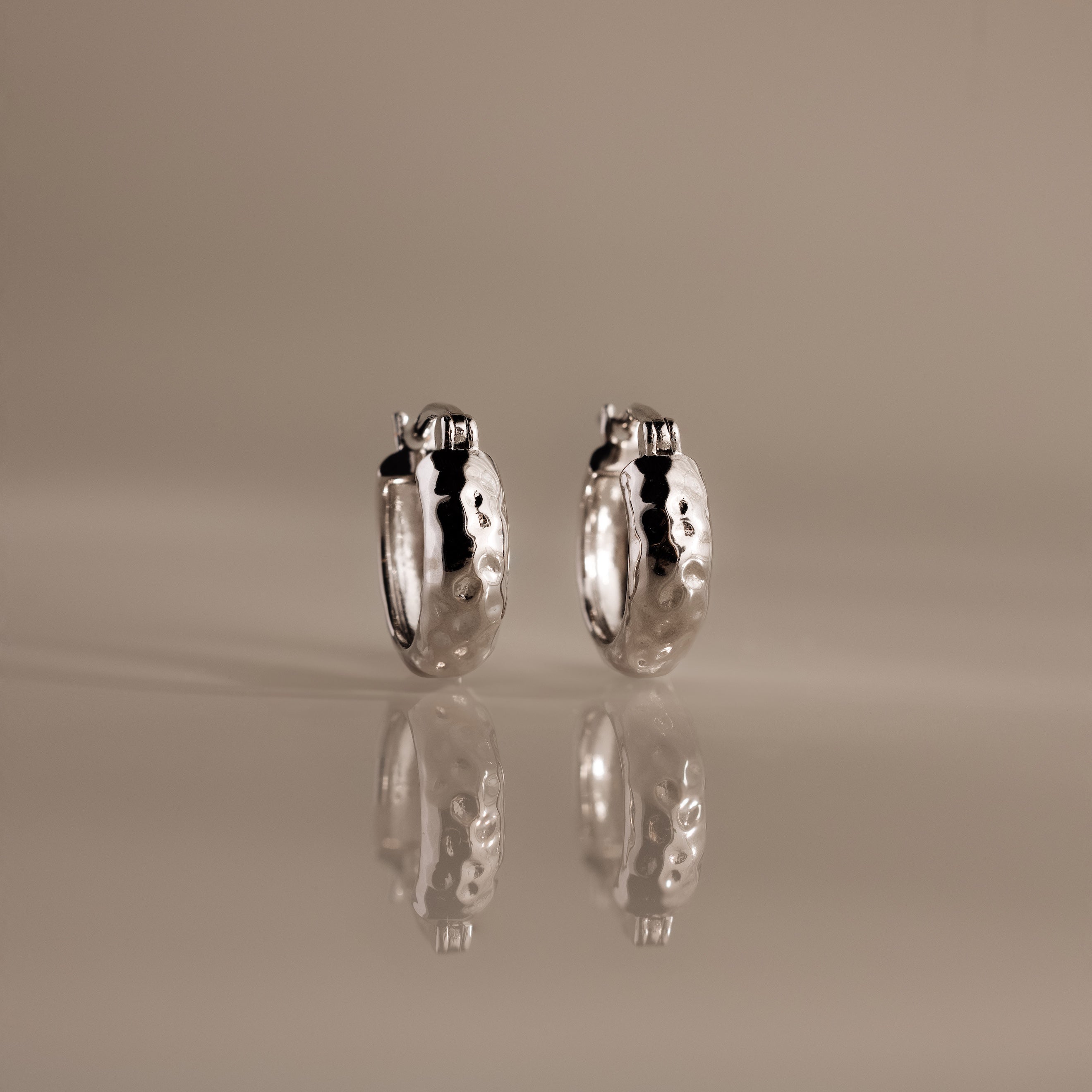 Bold Hammered Huggies in Silver with a textured finish stand upright on a reflective surface, highlighting their minimal design against a neutral background.