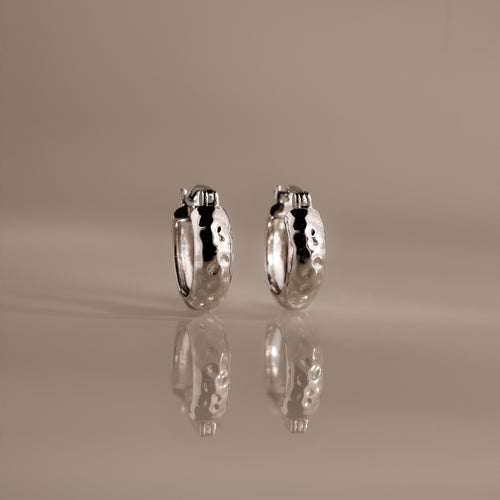 Bold Hammered Huggies in Silver with a textured finish stand upright on a reflective surface, highlighting their minimal design against a neutral background.