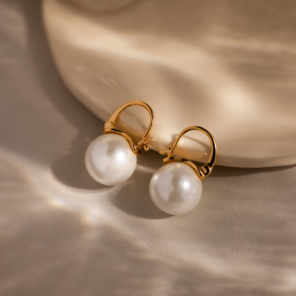 Gold hoop earrings with lustrous pearls rest on a light beige textured surface, highlighting the elegance of the Pearl Dome Drop Earrings in 18K Gold.