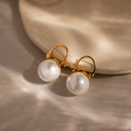 Gold hoop earrings with lustrous pearls rest on a light beige textured surface, highlighting the elegance of the Pearl Dome Drop Earrings in 18K Gold.