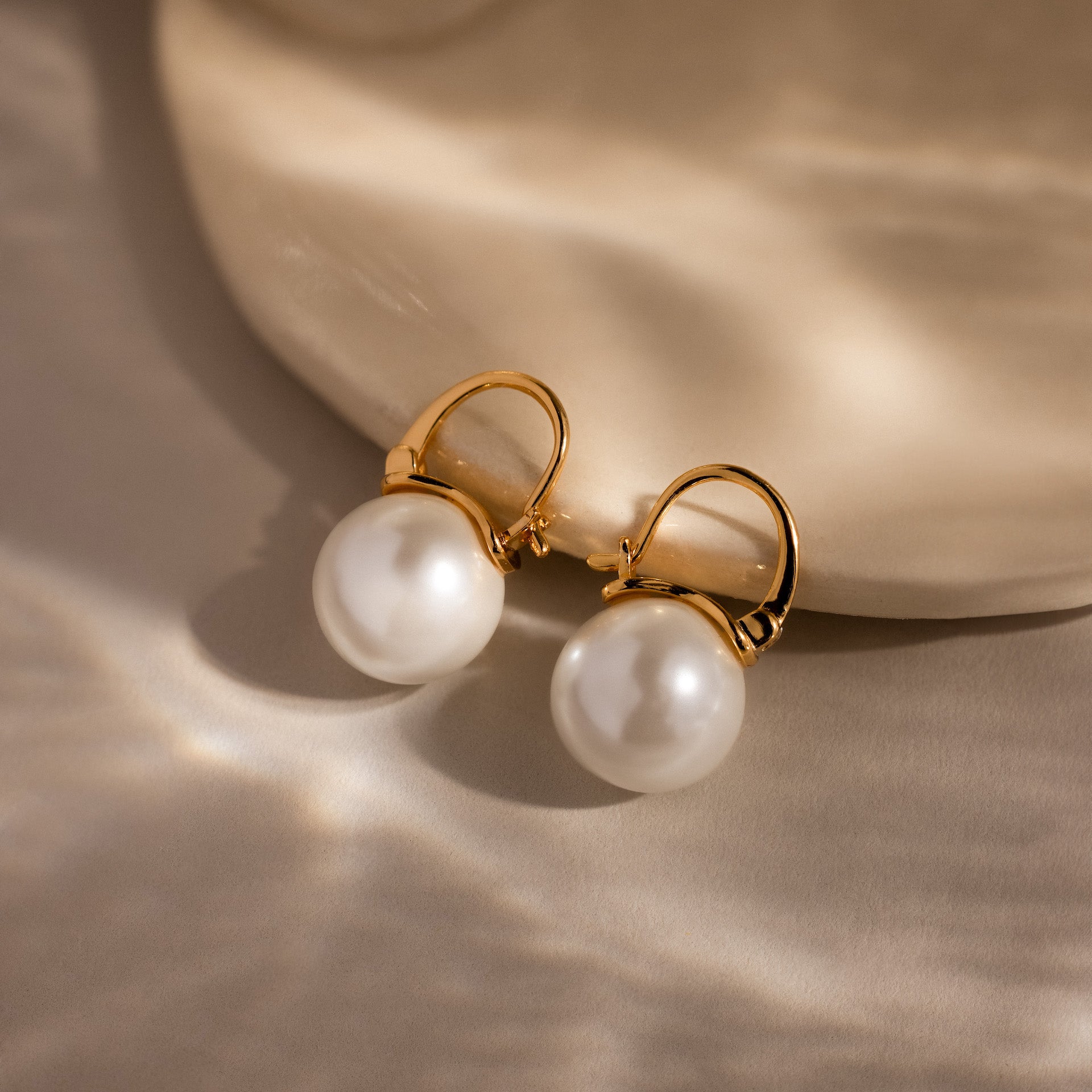 Gold hoop earrings with lustrous pearls rest on a light beige textured surface, highlighting the elegance of the Pearl Dome Drop Earrings in 18K Gold.
