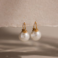 Pearl Dome Drop Earrings showcase lustrous pearls set against a textured beige background with soft shadows.