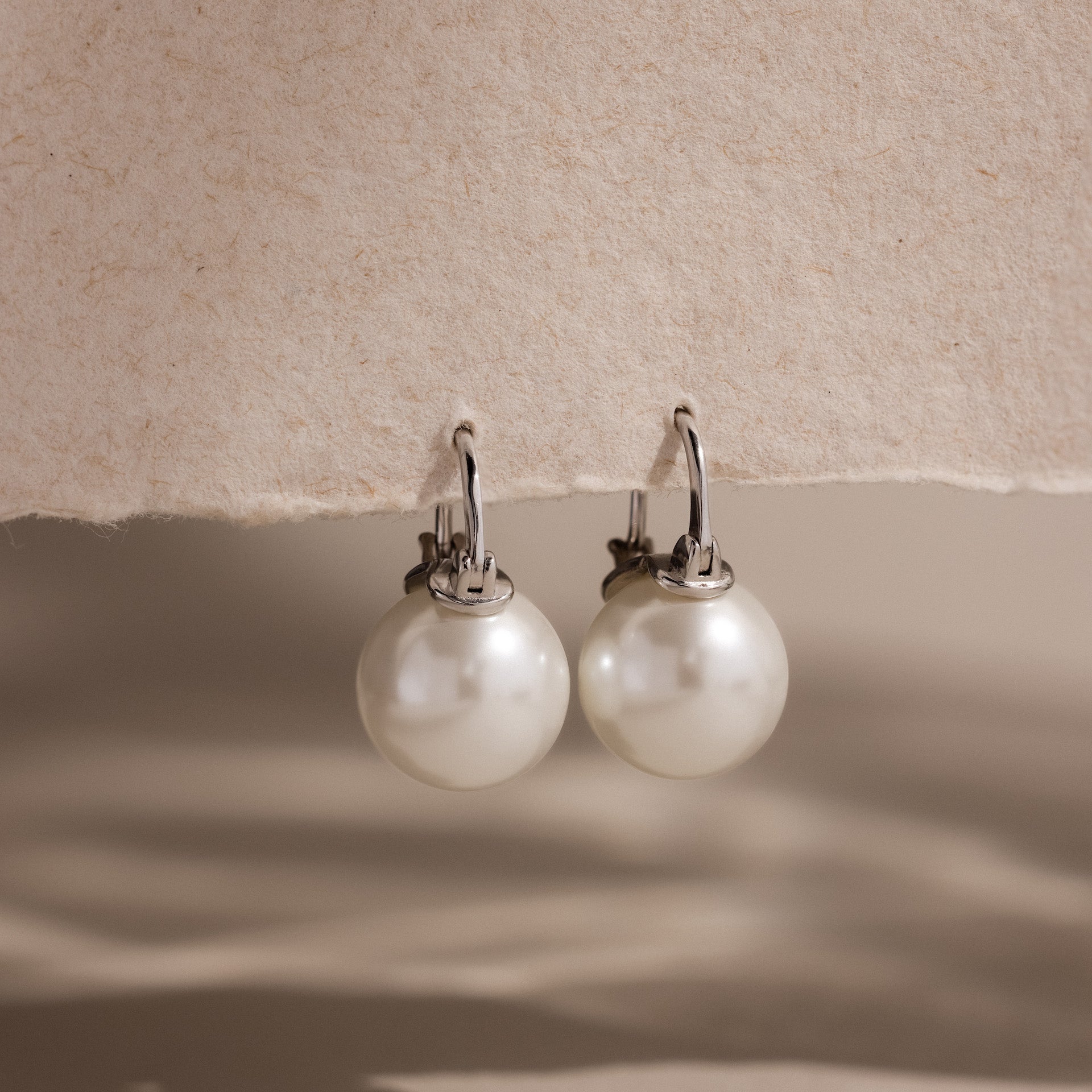 A pair of Pearl Dome Drop Earrings in Sterling Silver, featuring lustrous pearls and silver hooks, displayed on a textured beige surface.