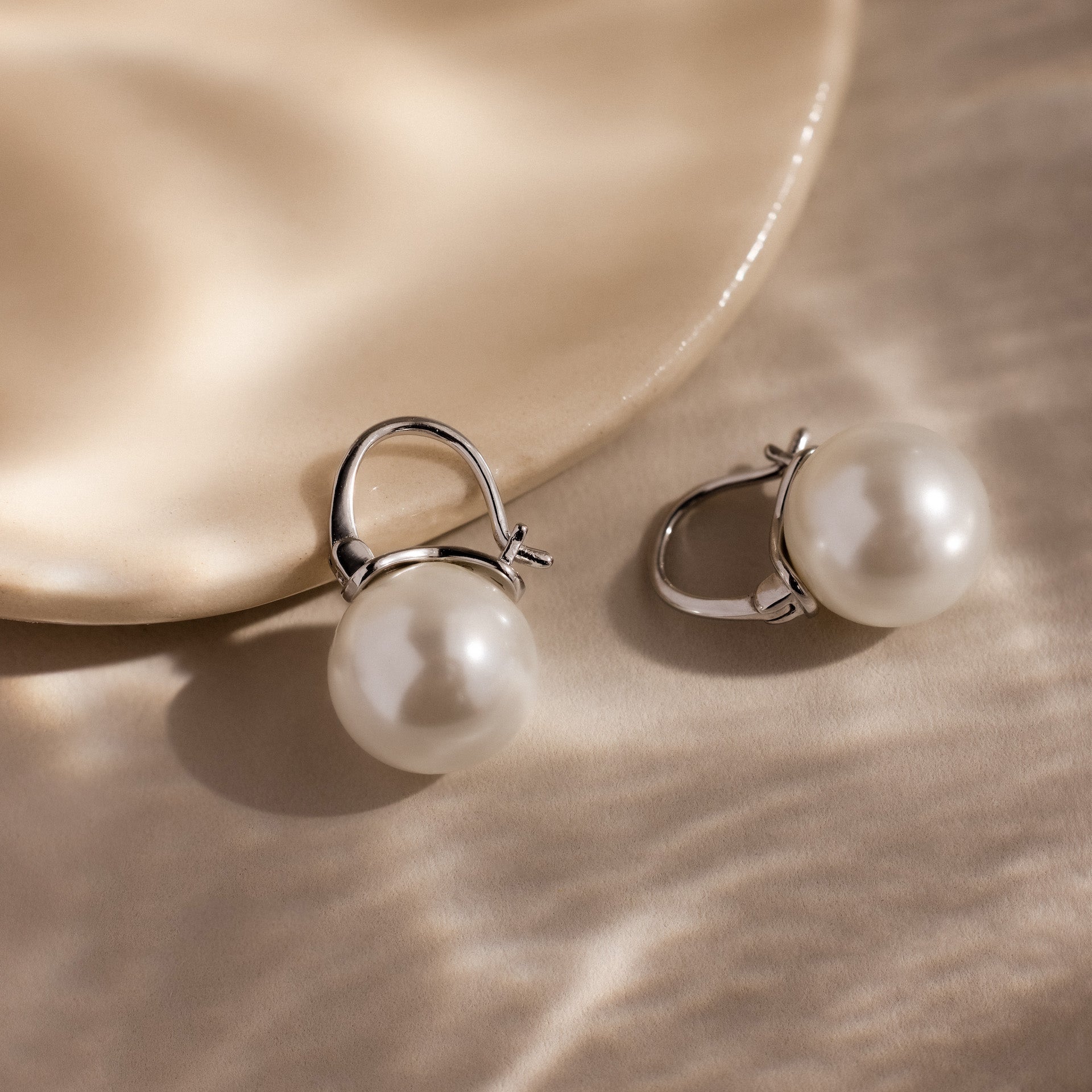 The Pearl Dome Drop Earrings, featuring lustrous pearls, are displayed on a softly lit beige surface.