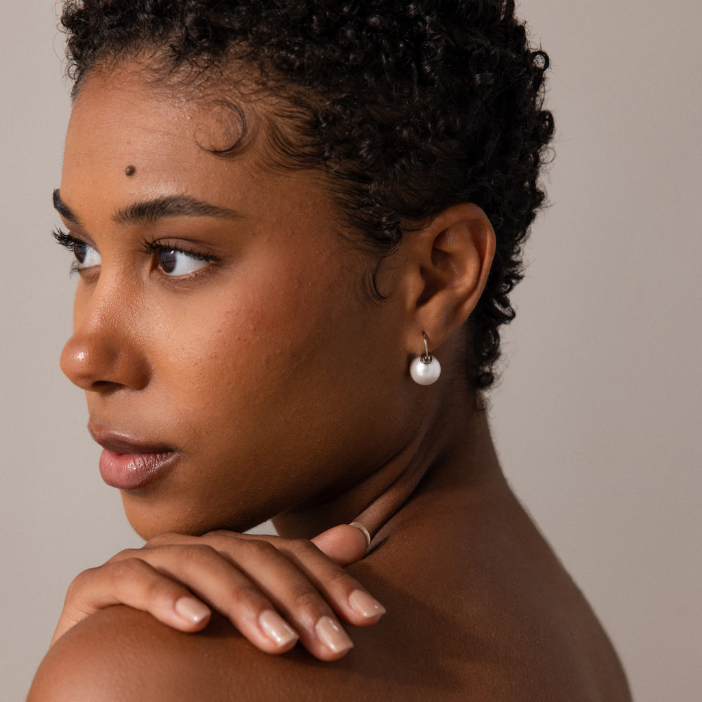 A woman with short, curly hair wears Pearl Dome Drop Earrings, gazing left with her hand on her shoulder against a neutral background.