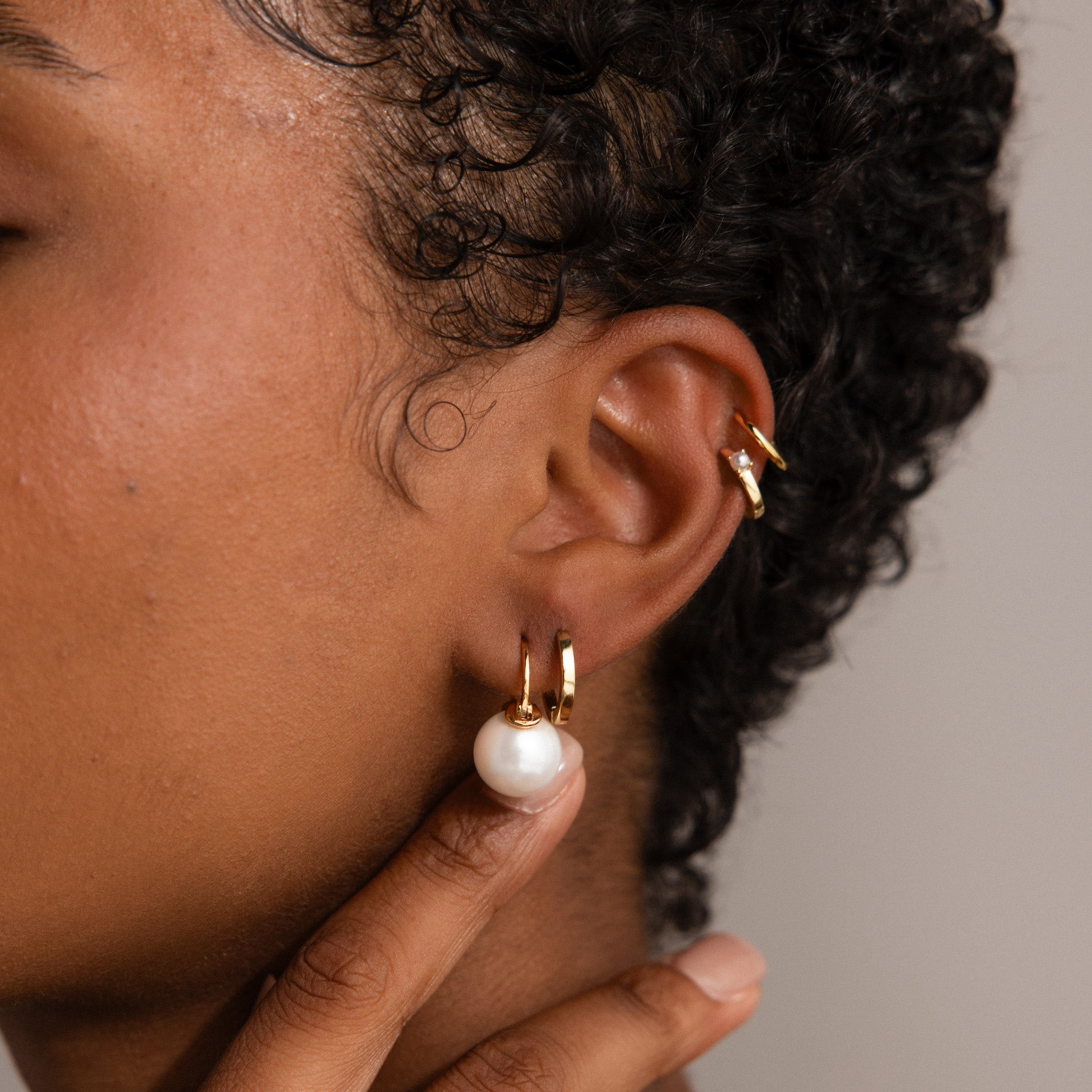 Close-up of a person wearing the Pearl Dome Drop Earrings in 18K Gold—gold hoops, one featuring a lustrous pearl—while gently touching their ear.