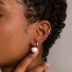 A close-up of an ear wearing elegant earrings, including a gold hoop and the Pearl Dome Drop Earrings with lustrous pearls, as a hand gently touches the earlobe.