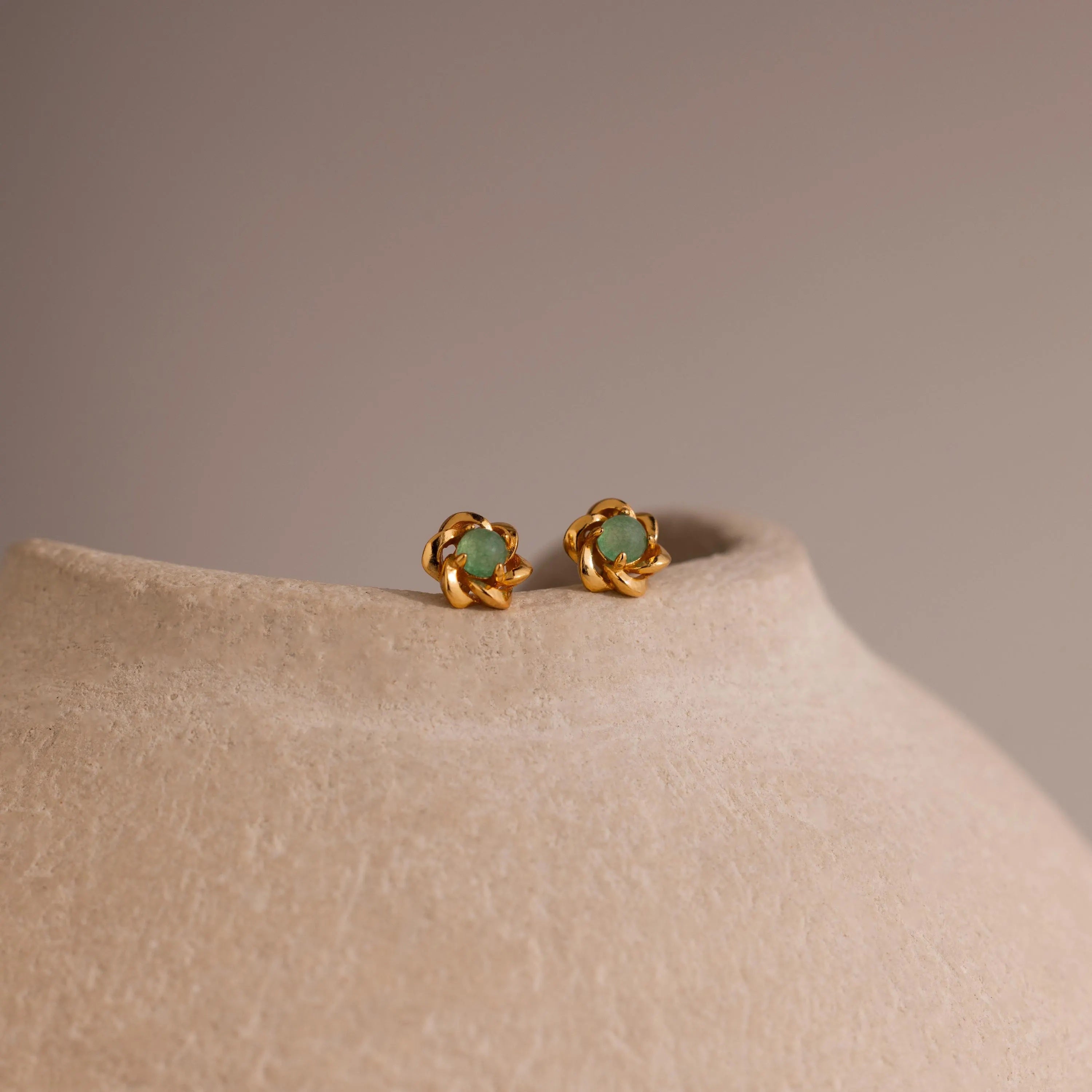 Close-up of gold flower-shaped stud earrings with a round green jade gemstone center, displayed on a neutral stone surface.