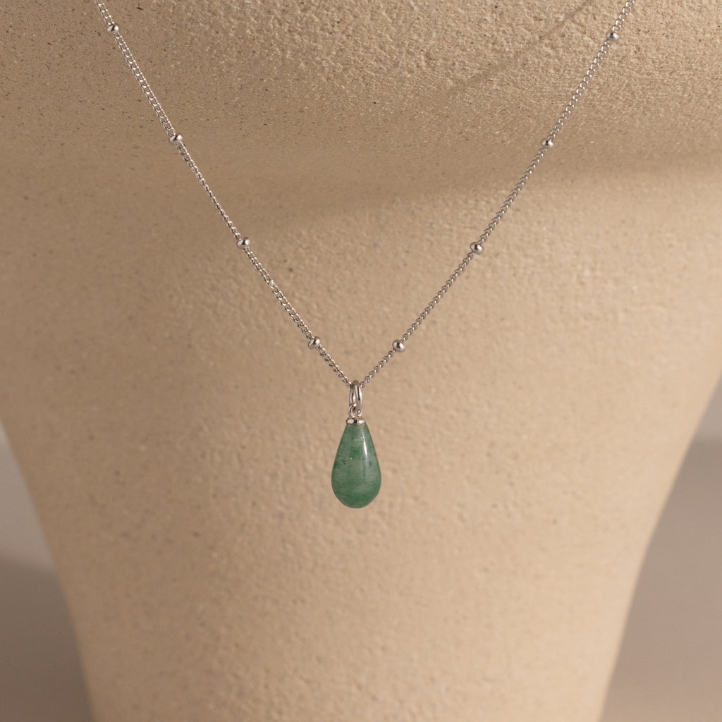 The Jade Drop Necklace features a silver beaded chain and a green teardrop jade pendant, presented on a beige jewelry stand.