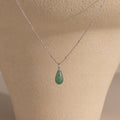 The Jade Drop Necklace features a silver beaded chain and a green teardrop jade pendant, presented on a beige jewelry stand.