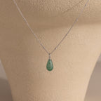 The Jade Drop Necklace features a silver beaded chain and a green teardrop jade pendant, presented on a beige jewelry stand.