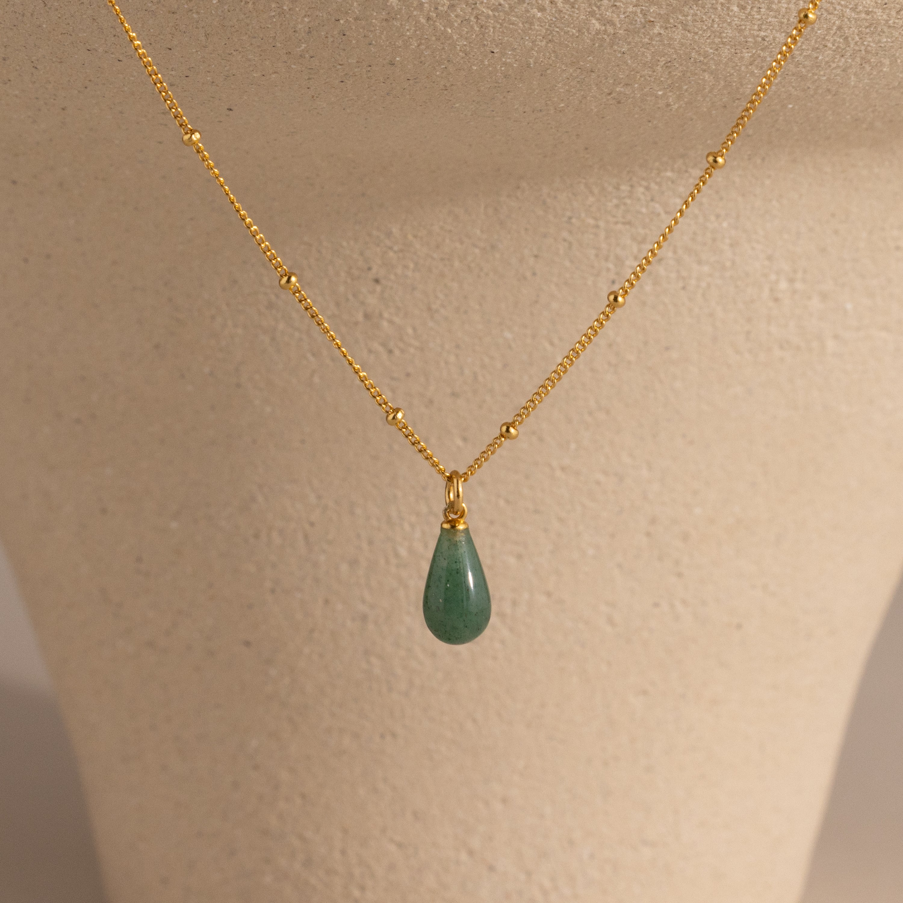 The Jade Drop Necklace, featuring a gold chain and green teardrop jade pendant, is displayed on a beige jewelry bust to highlight its elegant design.