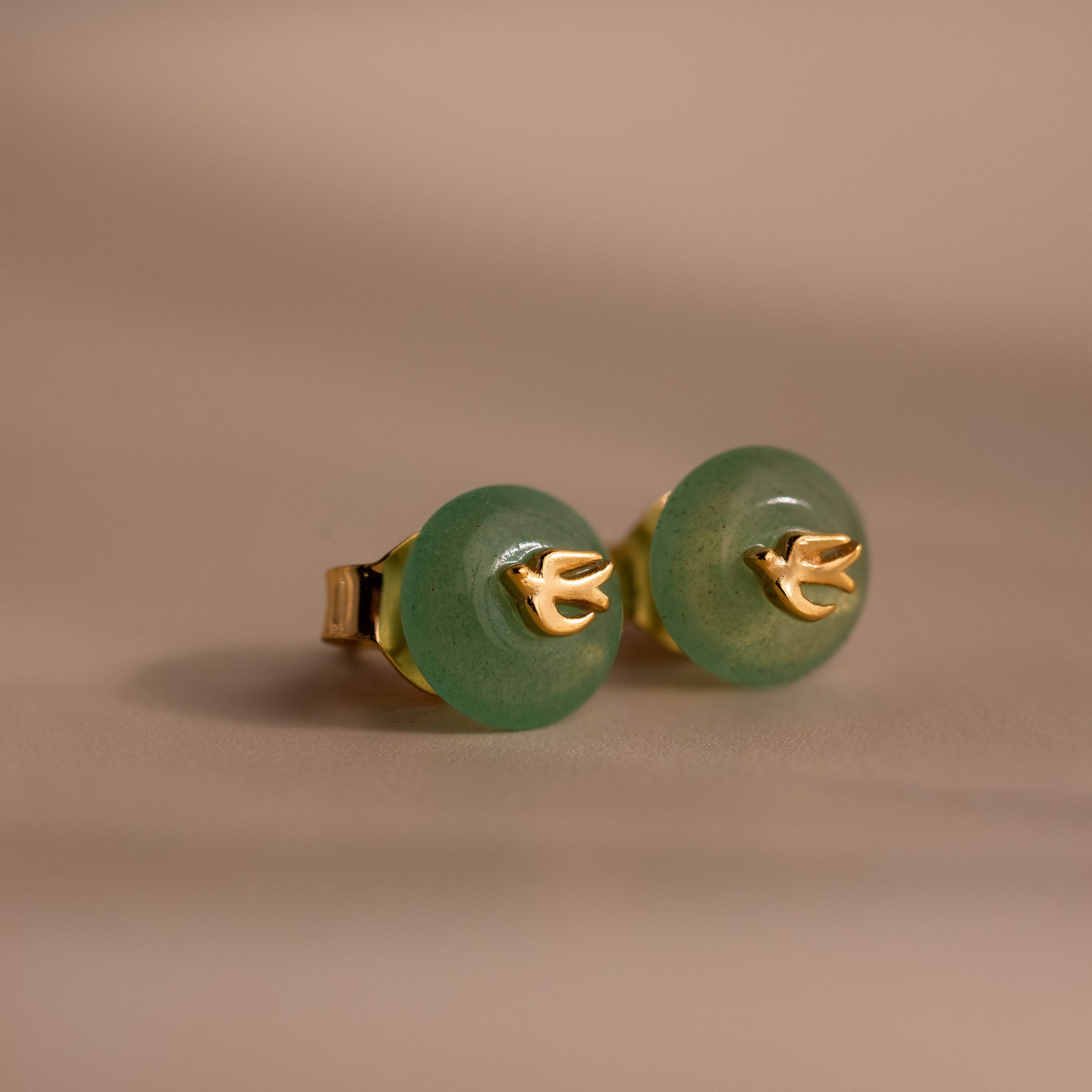 Close-up of gold Jade Bird Studs with dainty golden dove birds on round green jade discs, displayed on a neutral beige surface.