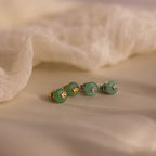 Two pairs of Jade Bird Studs in gold and silver, each with dainty dove birds on round green jade discs, displayed on a neutral beige surface.