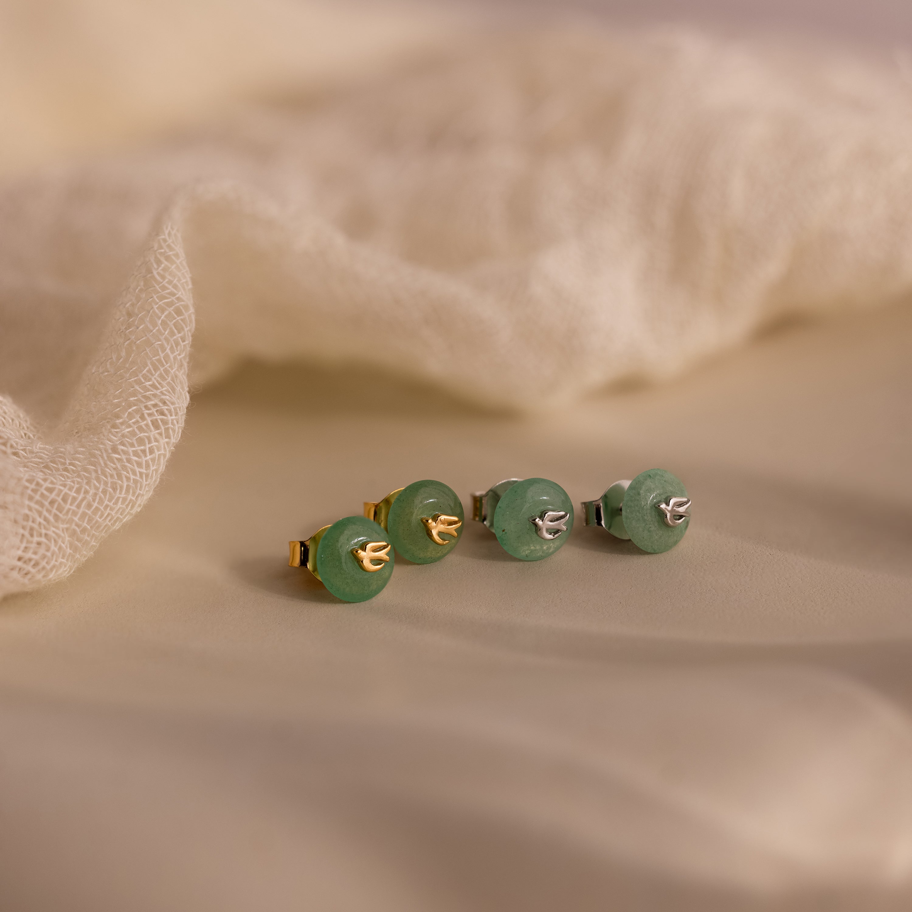 Two pairs of Jade Bird Studs in gold and silver, each with dainty dove birds on round green jade discs, displayed on a neutral beige surface.