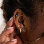 Model wearing a gold Jade Bird Stud Earring with a dainty golden dove bird on a round green jade disc, stacked with multiple gold and jade stud earrings.