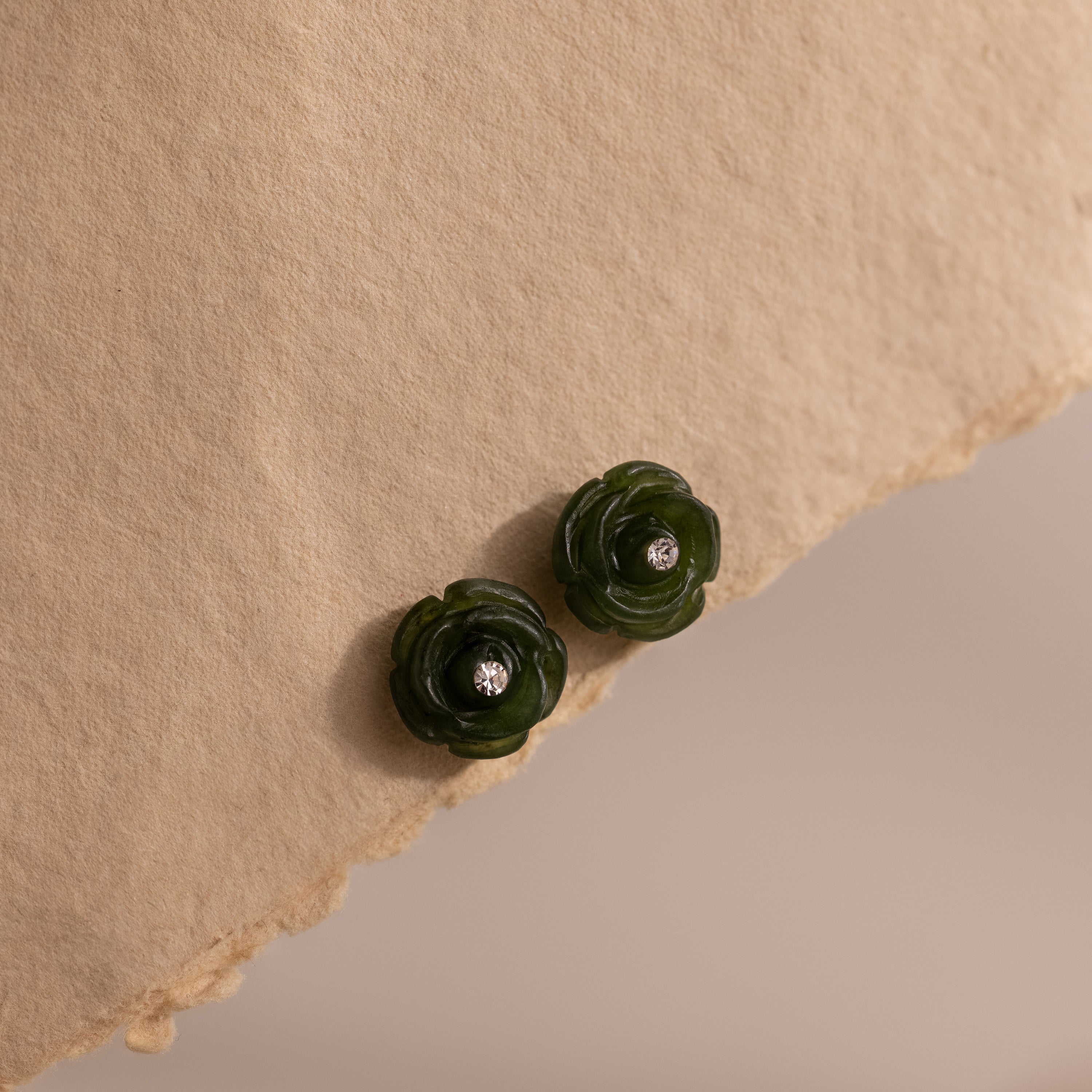 Pair of green jade rose earrings with faceted center stones displayed on a torn-edge neutral fabric background.