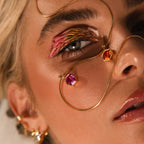 Close-up of a woman with bold eye makeup, gold geometric jewelry, and faceted stone pendants—featuring the Hexagon Pink Tourmaline Charm—draped across her skin.