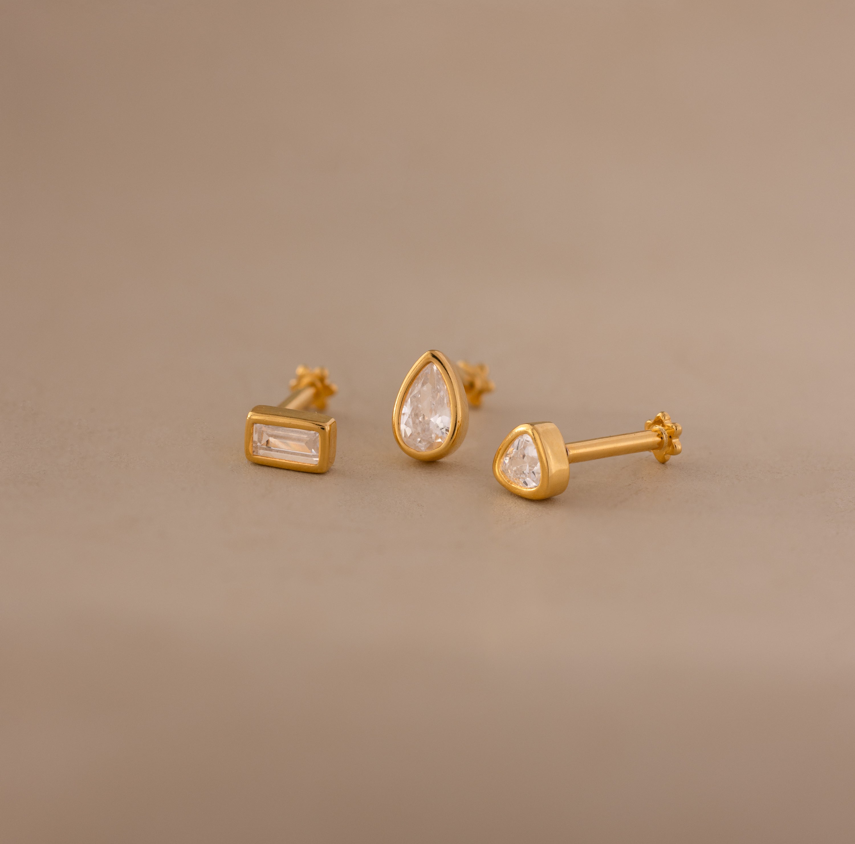 Three gold earrings from the Diamond Bezel Flatback Studs Set—rectangular, teardrop, and round—glimmer on a beige background, showcasing classic and chic designs for timeless elegance.