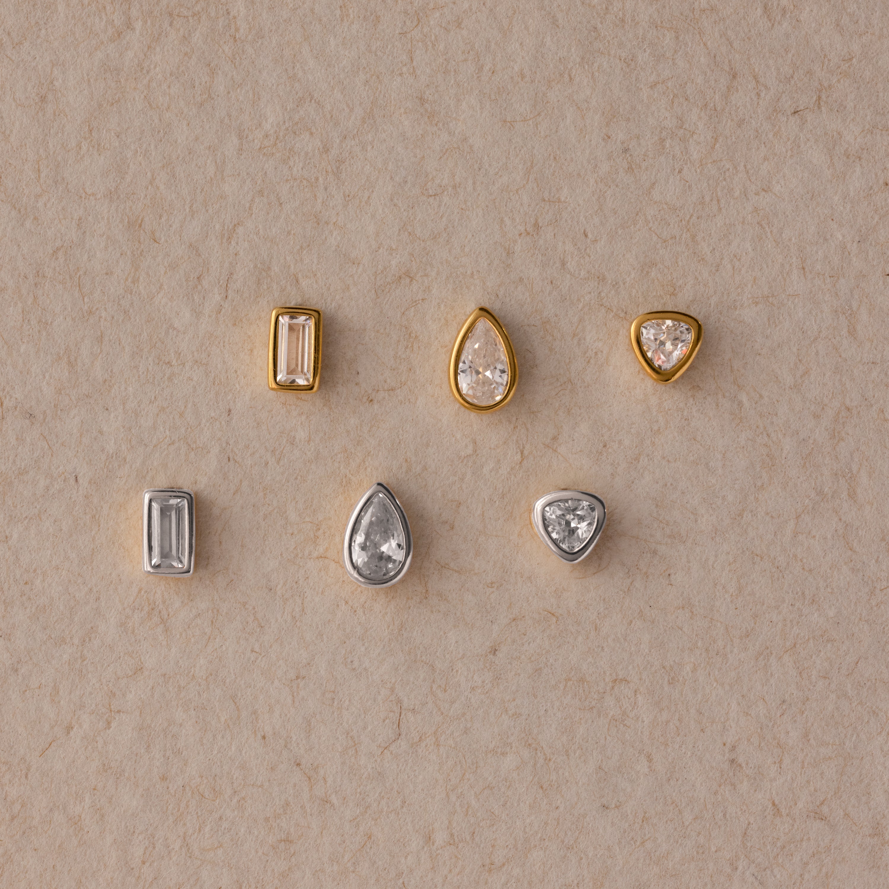 The Diamond Bezel Flatback Studs Set features six small gemstones in gold and silver settings, including classic stud earrings and diamond bezel studs, elegantly arranged on a beige surface.