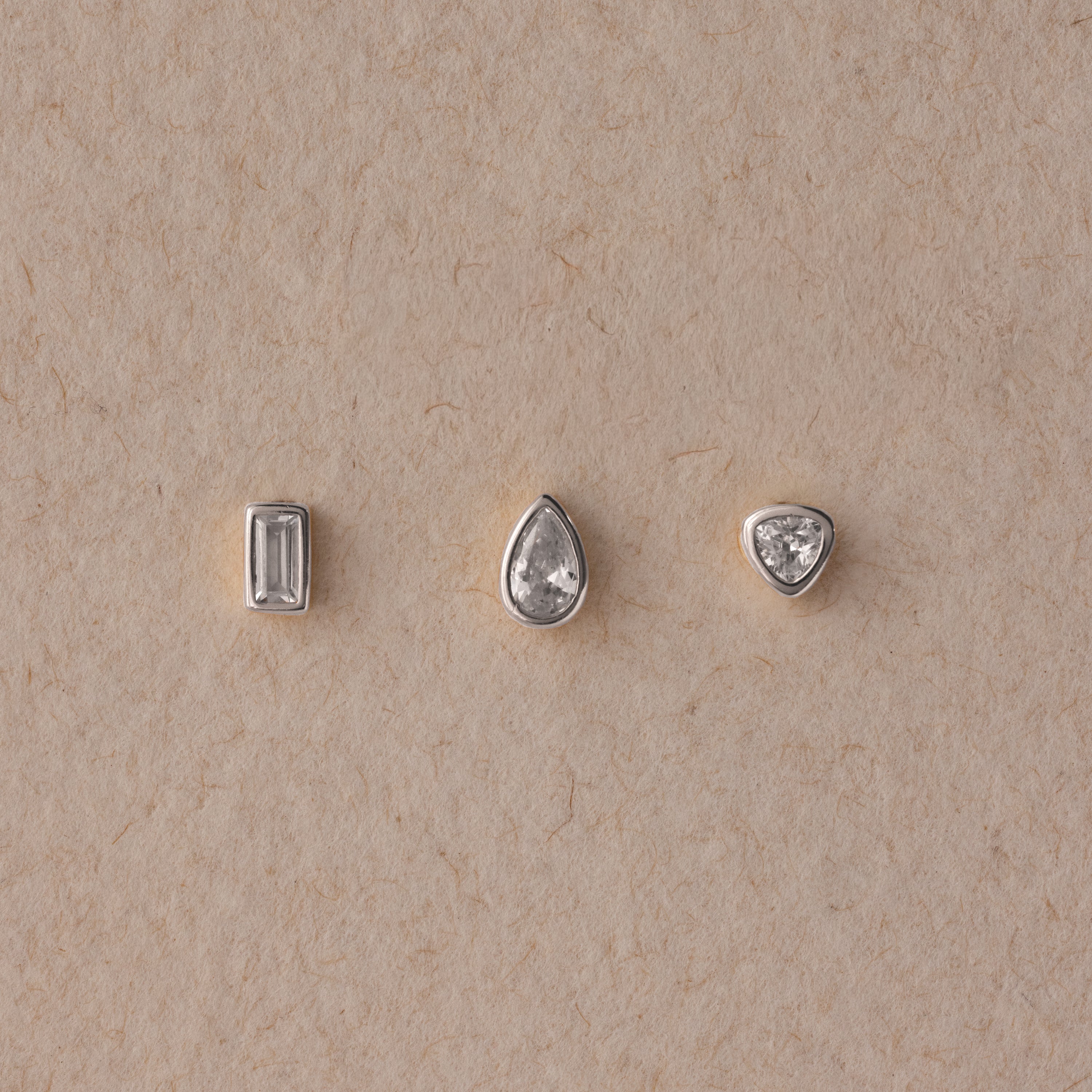 The Diamond Bezel Flatback Studs Set in Silver features rectangular, teardrop, and triangular gemstones on a beige textured background for a classic, elegant look.