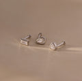 The Diamond Bezel Flatback Studs Set in Silver features three silver stud earrings with clear gemstones, including classic, modern, and bezel styles, displayed on a beige surface.