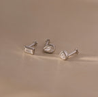 The Diamond Bezel Flatback Studs Set in Silver features three silver stud earrings with clear gemstones, including classic, modern, and bezel styles, displayed on a beige surface.