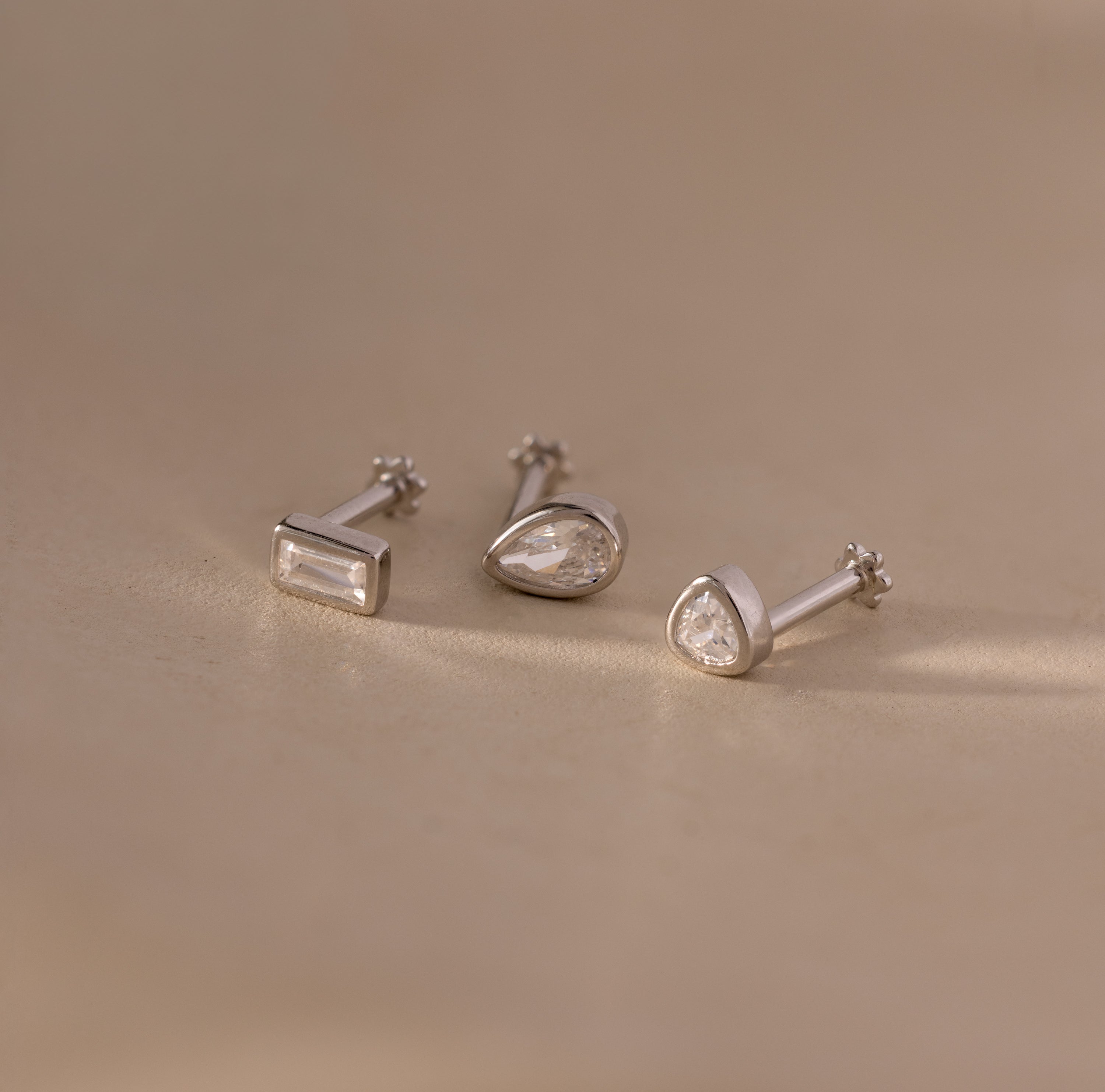 The Diamond Bezel Flatback Studs Set in Silver features three silver stud earrings with clear gemstones, including classic, modern, and bezel styles, displayed on a beige surface.