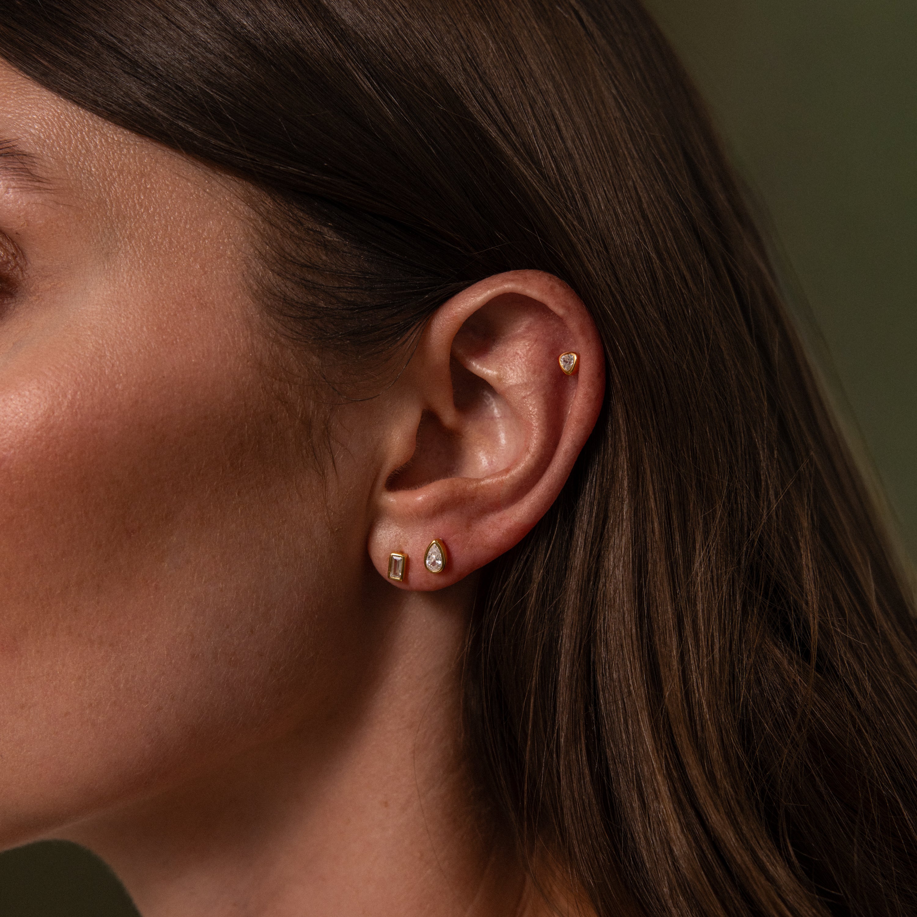 Close-up of a womans ear with the Diamond Bezel Flatback Studs Set, featuring three gold earrings—heart-shaped stud, two small hoops, and classic studs—for a timeless look.