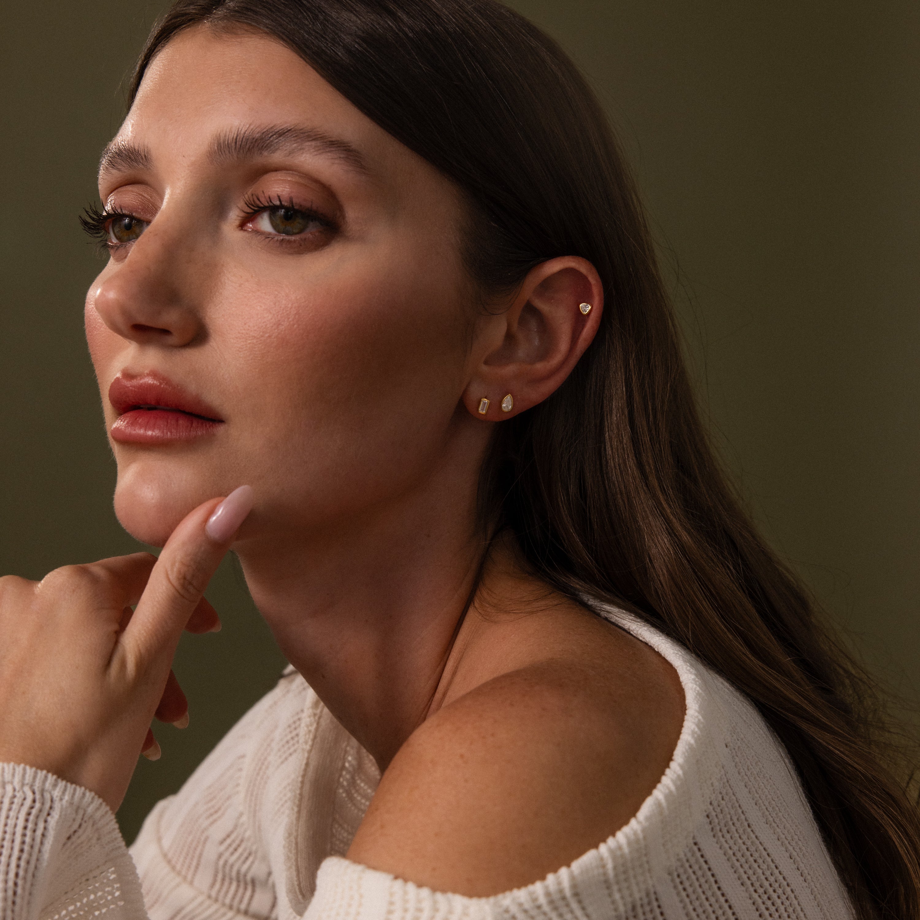 Woman with long brown hair wearing Diamond Bezel Flatback Studs Set and a white sweater, gazing thoughtfully to the side.