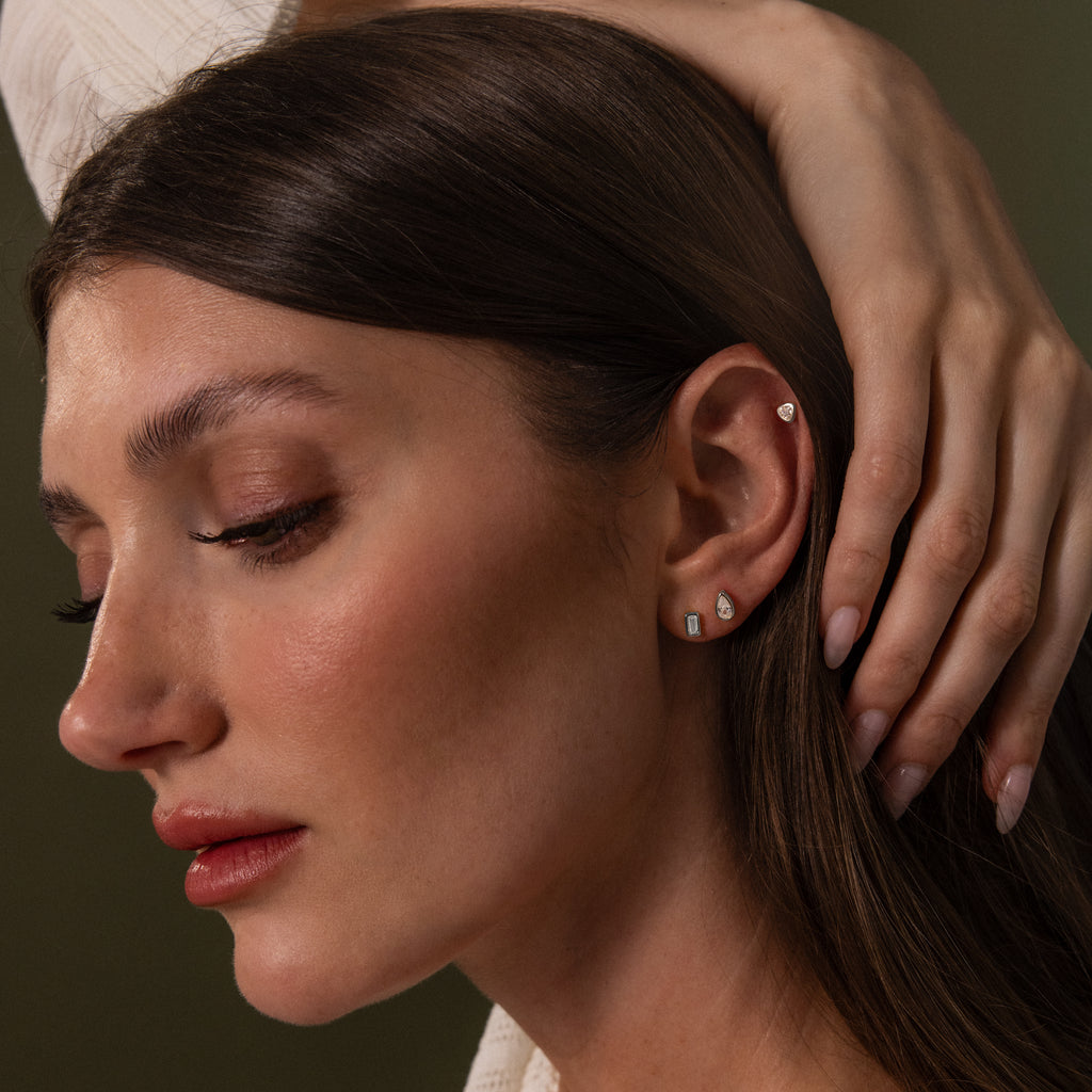 A woman with long brown hair touches her head, revealing three small earrings on her ear, including the elegant Diamond Bezel Flatback Studs Set.