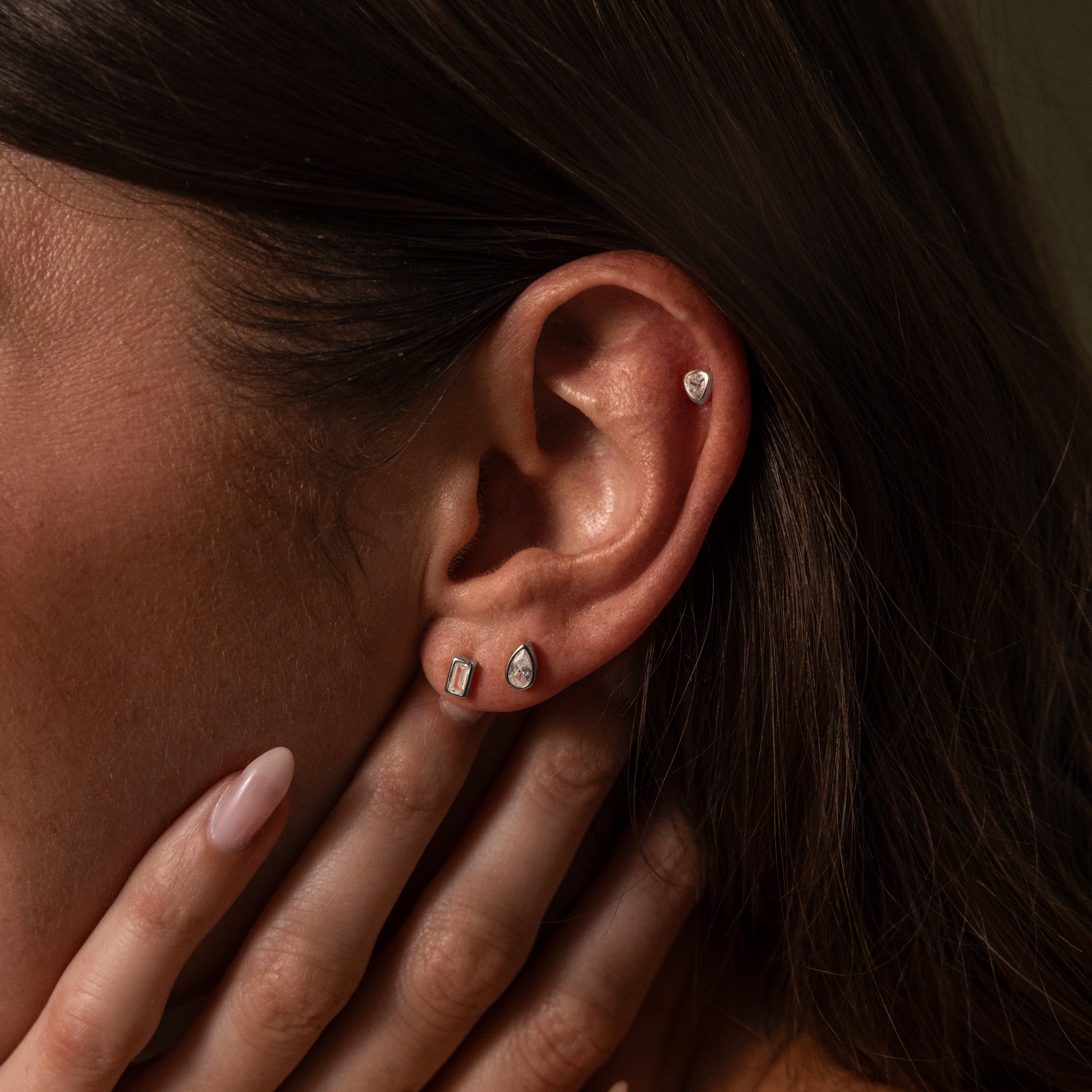 A close-up of a womans ear wearing three silver earrings, including Diamond Bezel Flatback Studs, as her hand gently touches her face.
