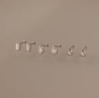 The Diamond Bezel Flatback Studs Set in Silver features three pairs of classic cut earrings—rectangular, teardrop, and round—elegantly displayed on a beige background.
