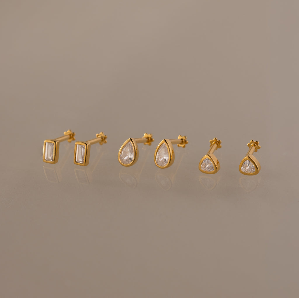 The Diamond Bezel Flatback Studs Set features three pairs of classic stud earrings—rectangular, teardrop, and round clear gemstones—shown on a neutral background.