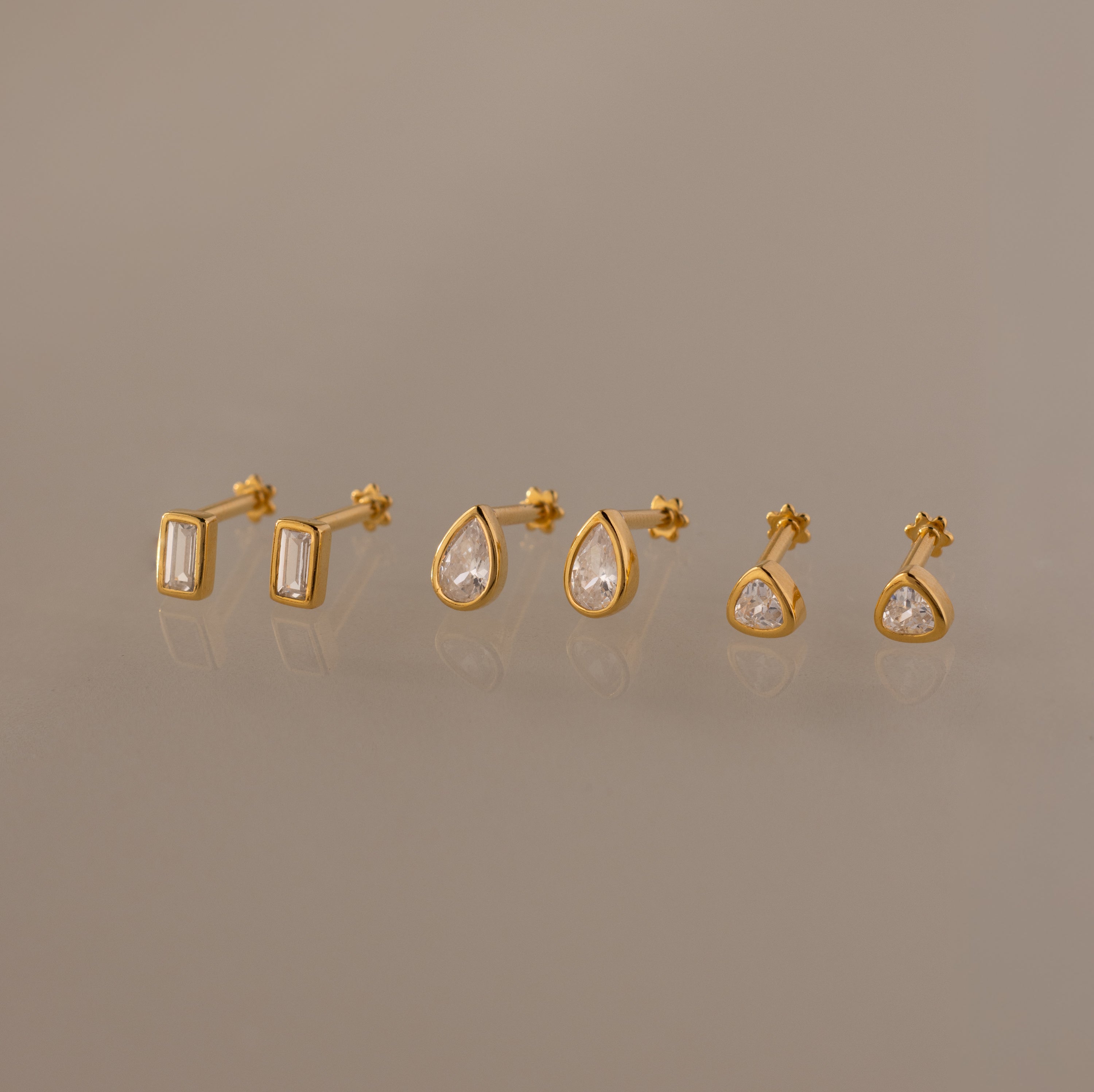 The Diamond Bezel Flatback Studs Set features three pairs of classic stud earrings—rectangular, teardrop, and round clear gemstones—shown on a neutral background.